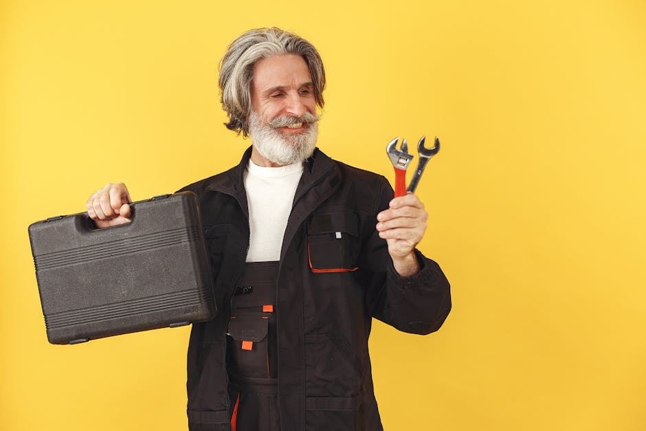 Elderly bearded man with safety glasses smiling, holding a toolbox and wrenches against a yellow background.