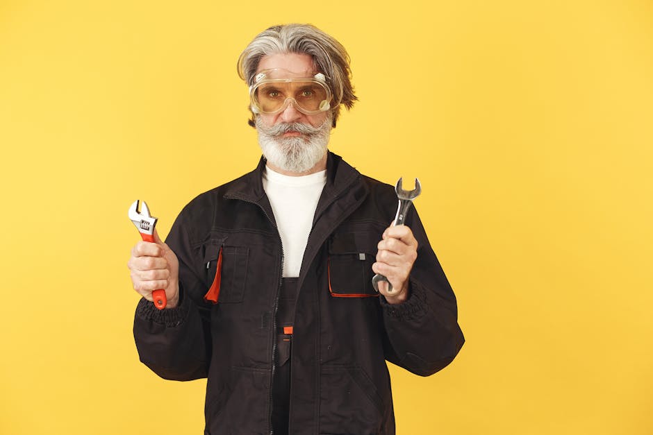 Senior man in black workwear holding wrenches, standing against a bright yellow background.